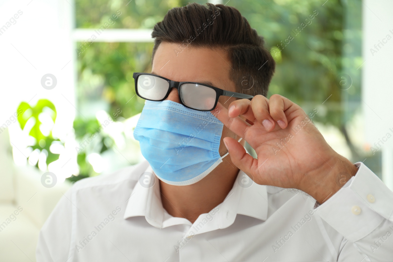 Photo of Man wiping foggy glasses caused by wearing medical mask indoors, closeup