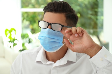 Man wiping foggy glasses caused by wearing medical mask indoors, closeup