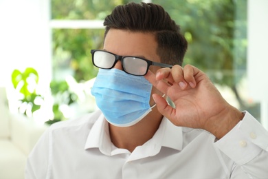 Photo of Man wiping foggy glasses caused by wearing medical mask indoors, closeup