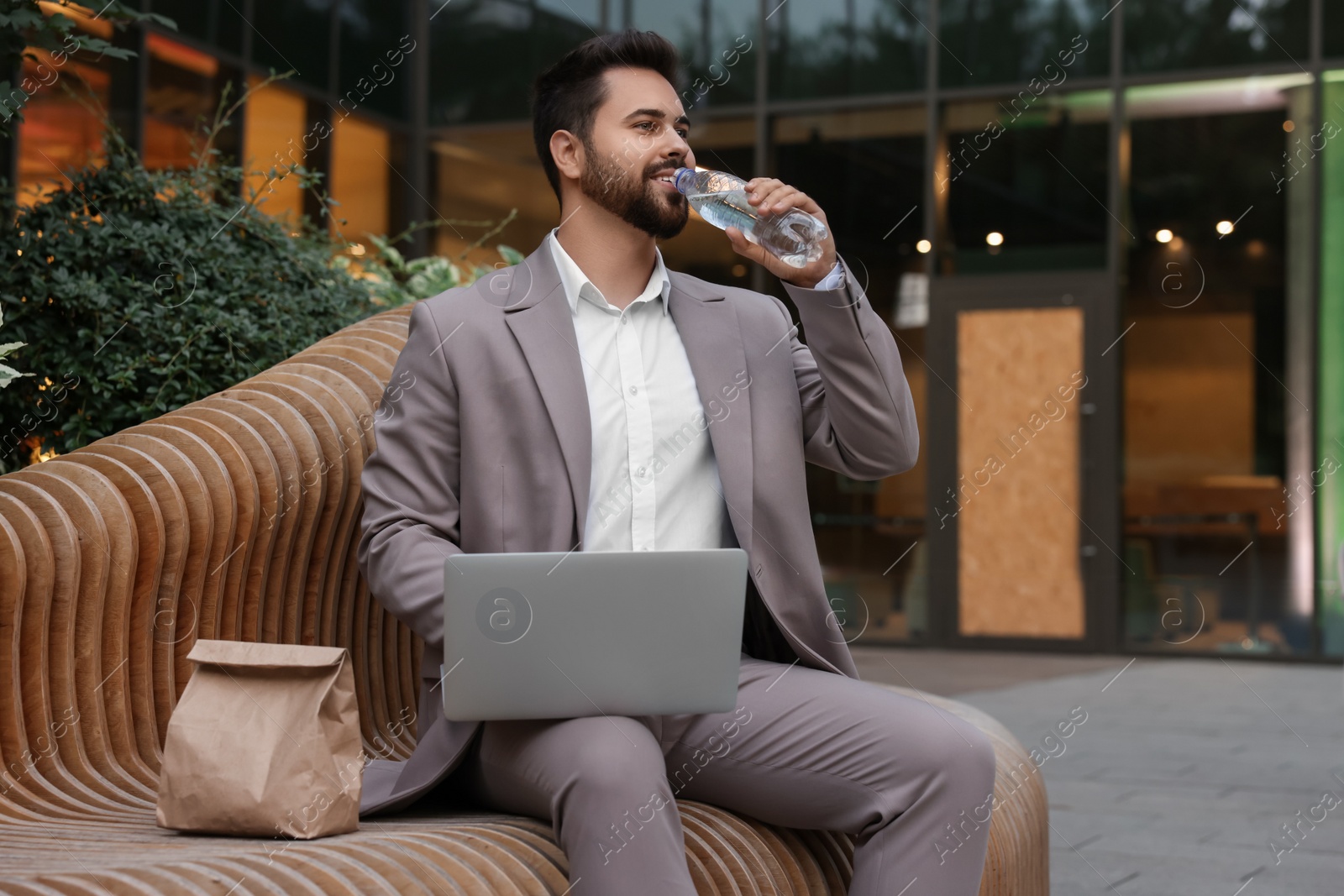 Photo of Young businessman with laptop drinking water from bottle while lunch on bench outdoors