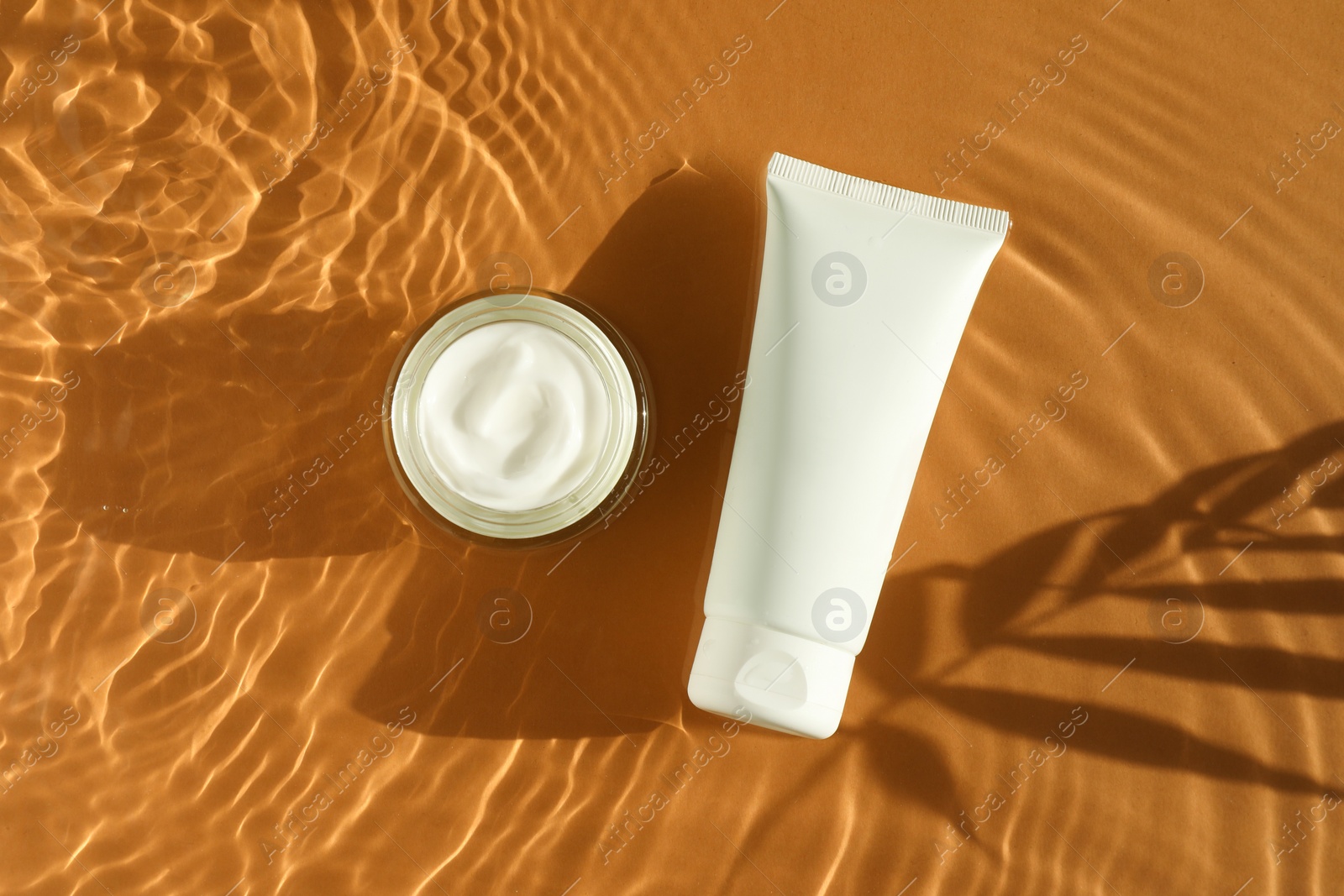 Photo of Tube and jar with moisturizing cream in water on orange background, top view