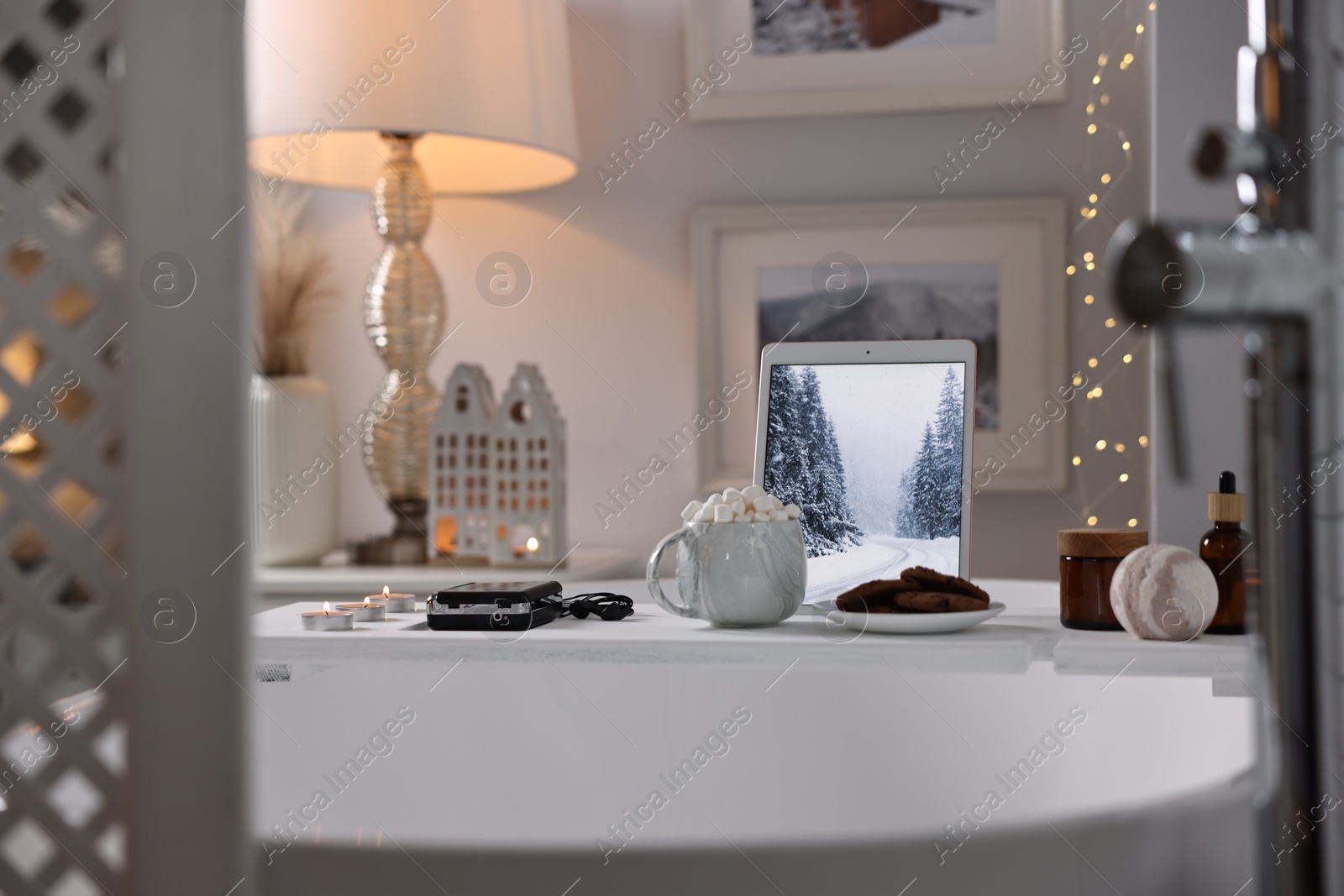 Photo of White wooden tray with tablet, spa products and burning candles on bathtub in bathroom