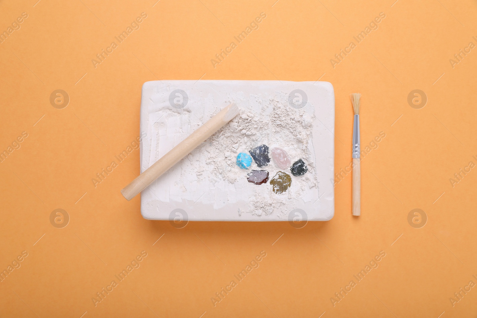 Photo of Educational toy for motor skills development. Excavation kit (plaster, wooden chisel, brush and gemstones) on orange background, flat lay