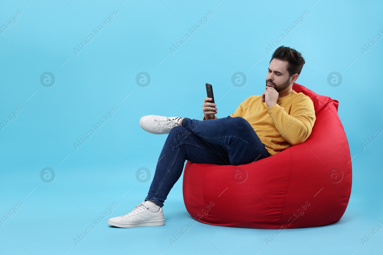 Photo of Handsome young man using smartphone on bean bag chair against light blue background. Space for text