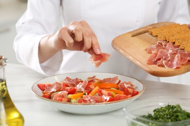 Professional chef adding prosciutto into delicious dish at white marble table, closeup