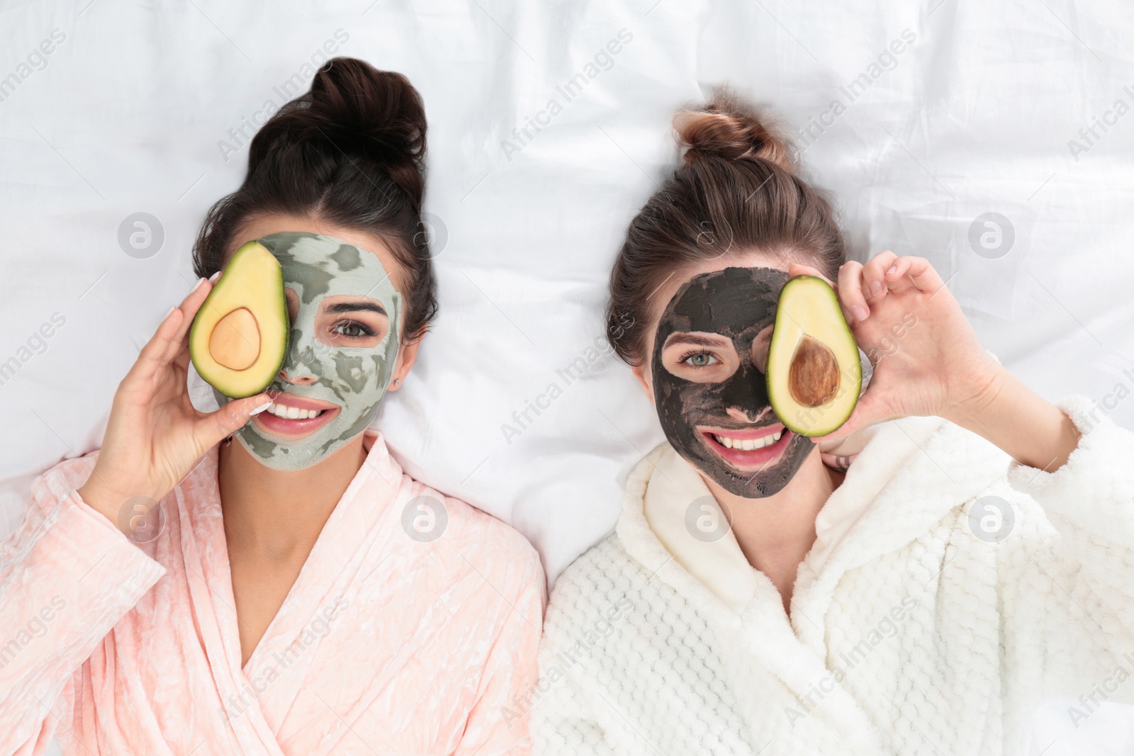 Photo of Young friends with facial masks having fun on bed at pamper party, top view