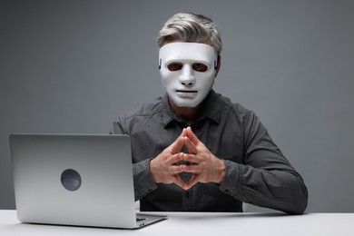 Photo of Man in mask sitting at white table with laptop against grey background