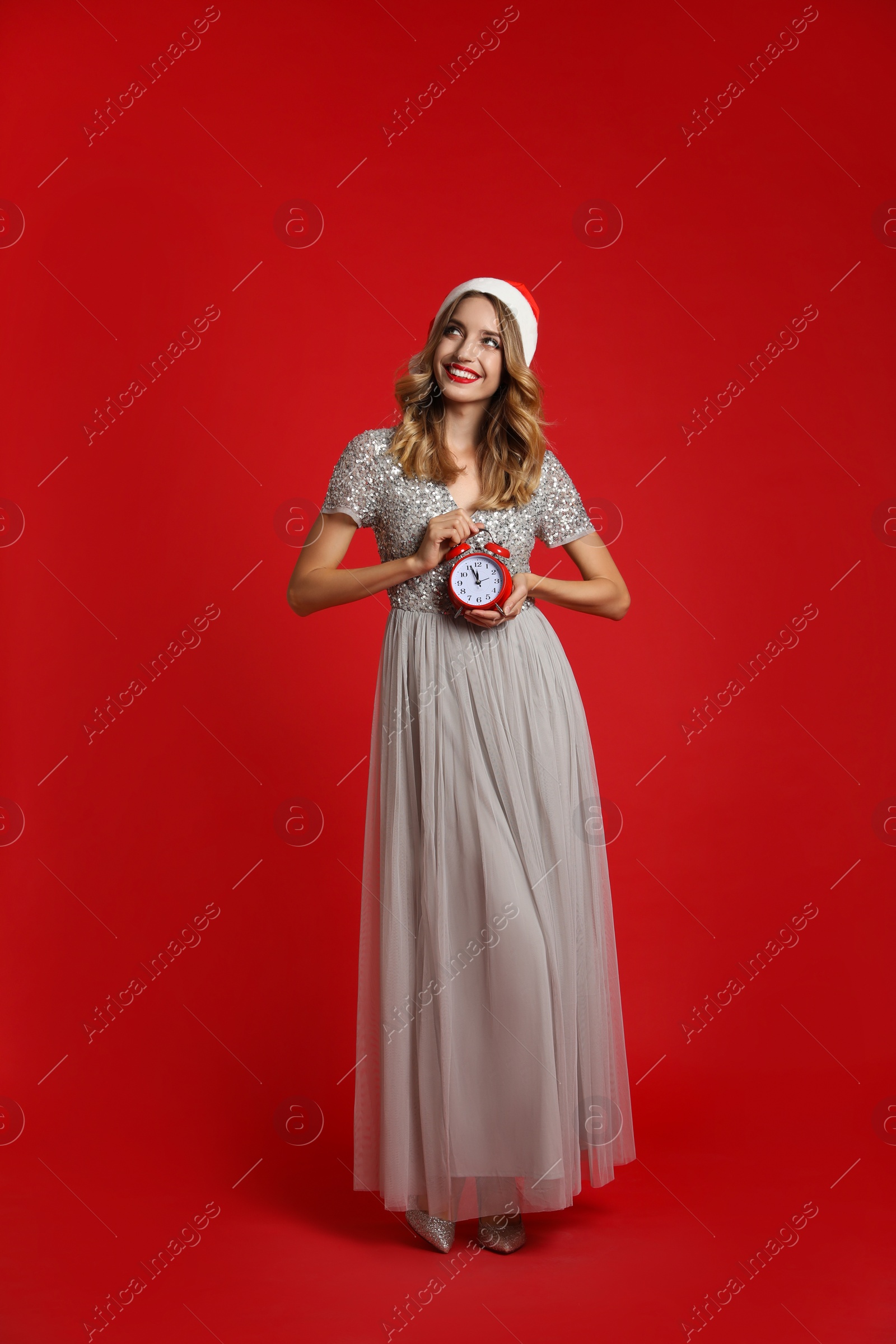 Photo of Happy young woman wearing Santa hat with alarm clock on red background. Christmas time