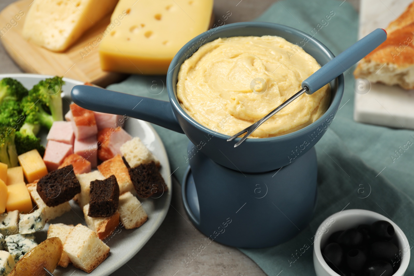 Photo of Tasty melted cheese in fondue pot, fork and snacks on grey table, closeup