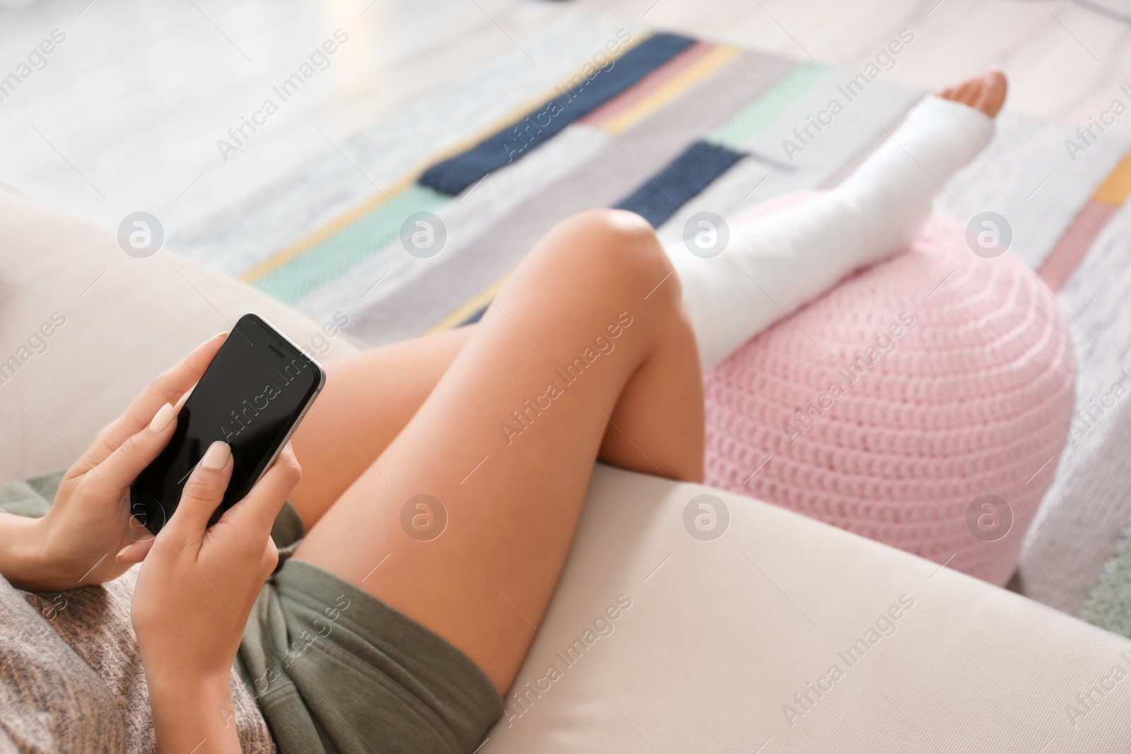 Photo of Young woman with broken leg in cast using mobile phone while sitting on sofa at home