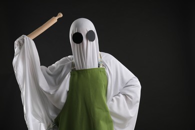 Woman in ghost costume and apron with rolling pin on black background, space for text