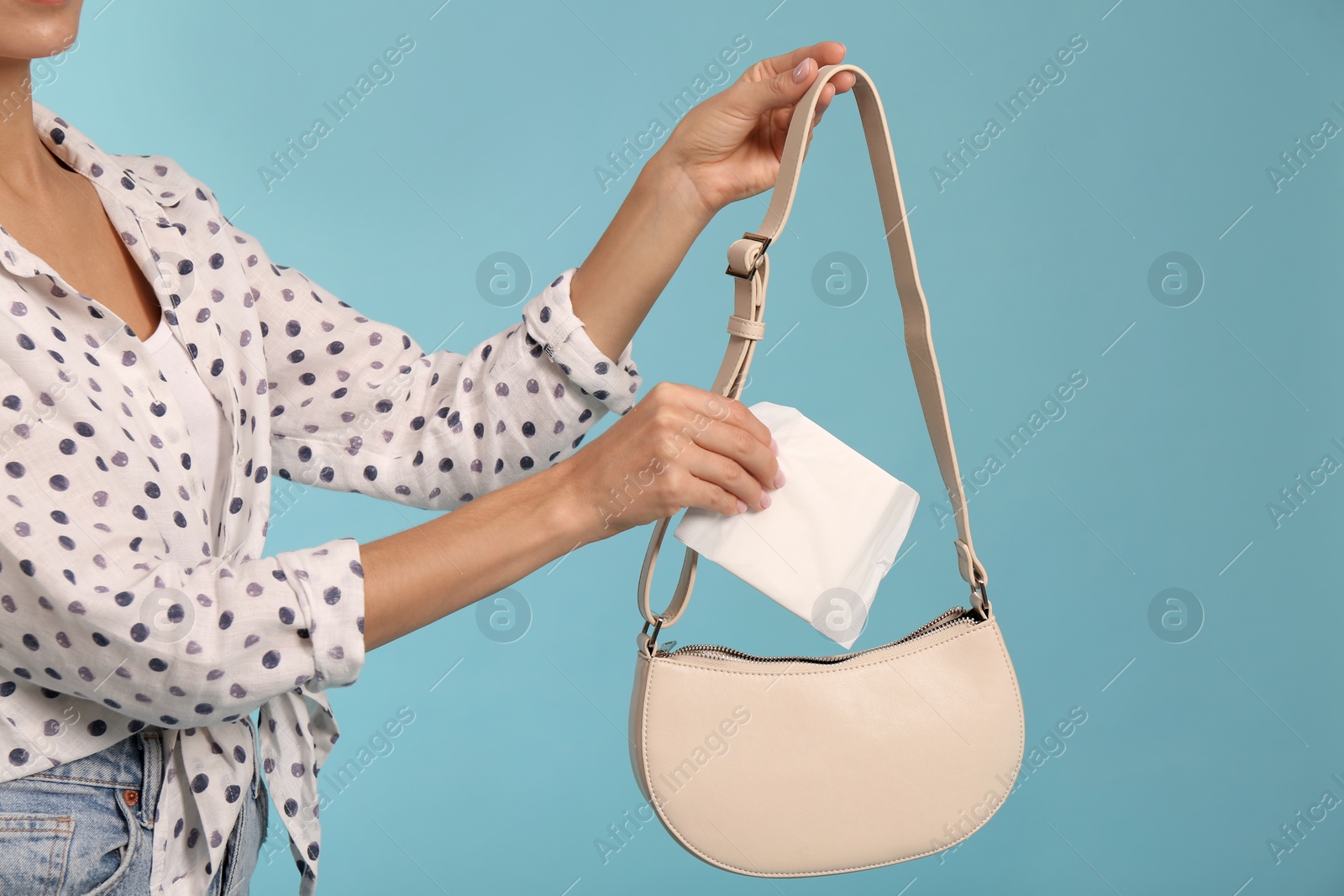 Photo of Young woman putting menstrual pad into bag on light blue background, closeup