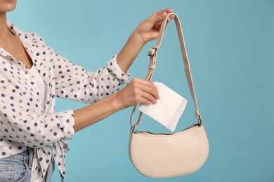 Young woman putting menstrual pad into bag on light blue background, closeup