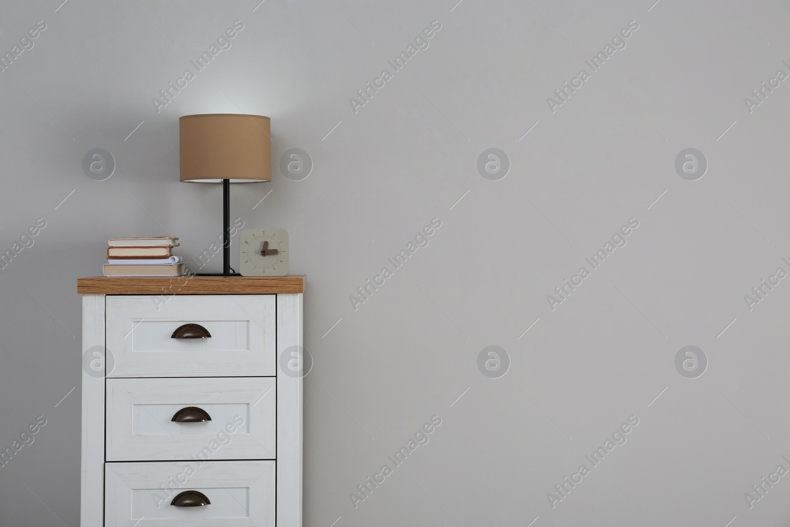 Photo of Chest of drawers with lamp, books and clock near light wall, space for text. Interior design