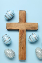 Wooden cross and painted Easter eggs on light blue background, flat lay