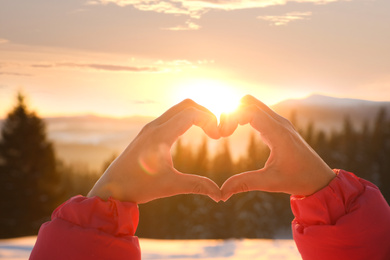 Photo of Woman making heart with hands outdoors at sunset, closeup. Winter vacation