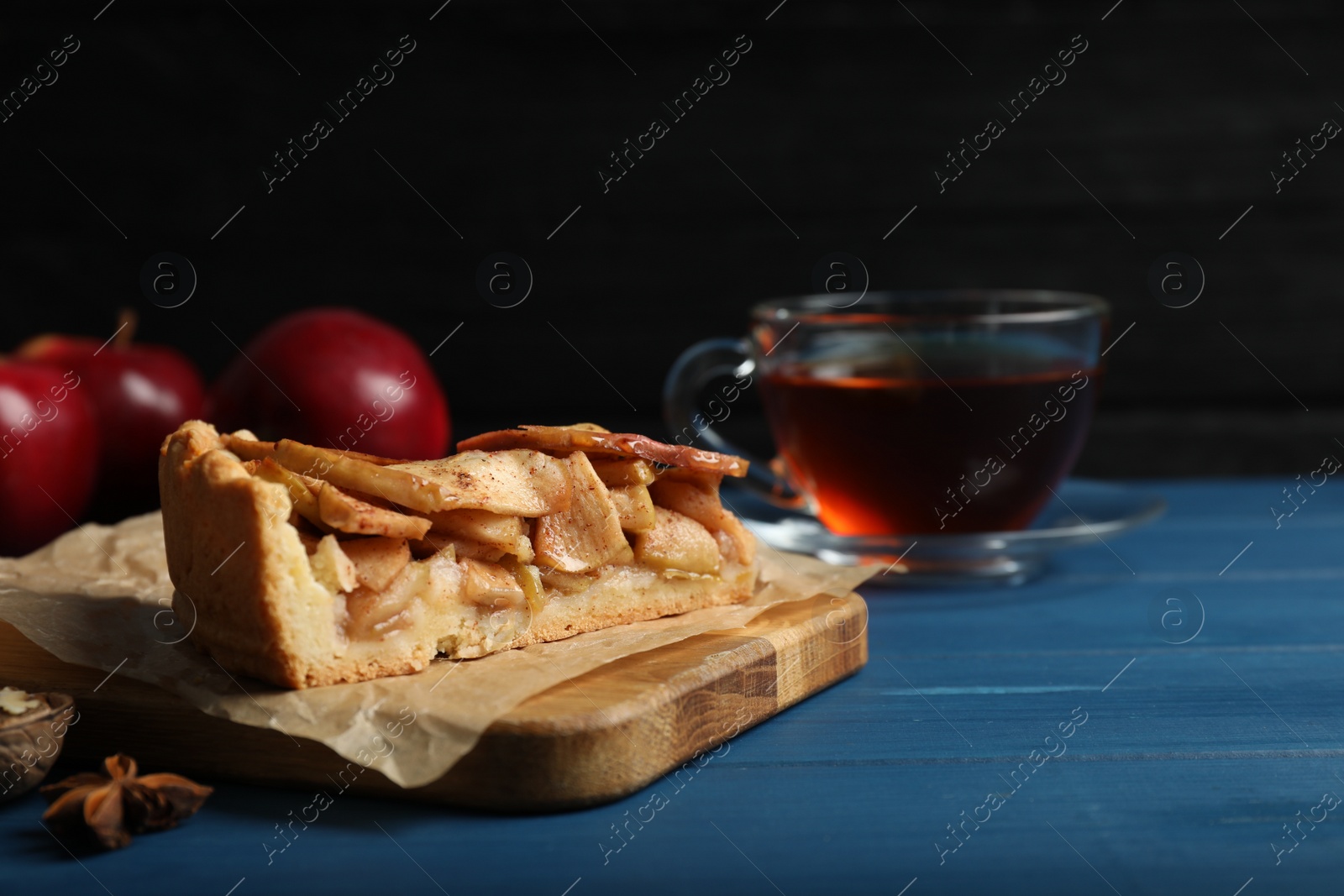 Photo of Slice of delicious apple pie served with tea on blue wooden table against dark background