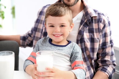Cute little boy with glass of milk sitting on father's lap indoors