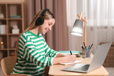 Photo of Online learning. Smiling teenage girl writing in notebook near laptop at home