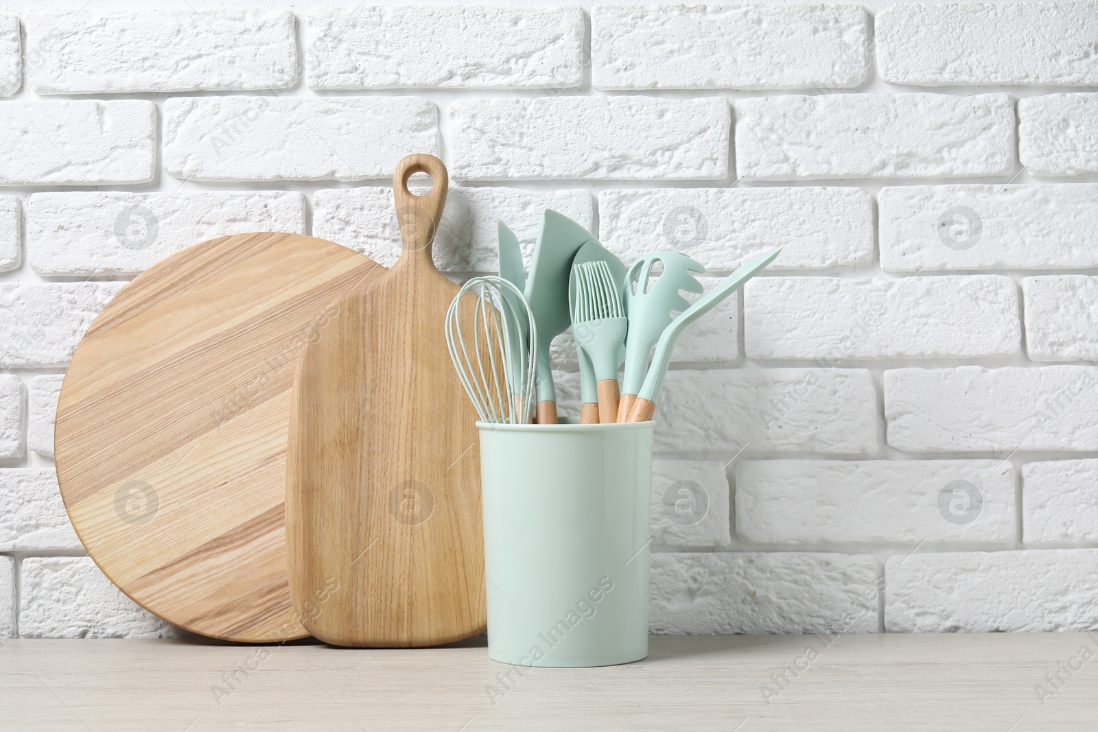 Photo of Different cutting boards and kitchen utensils in holder on light table near white brick wall. Space for text