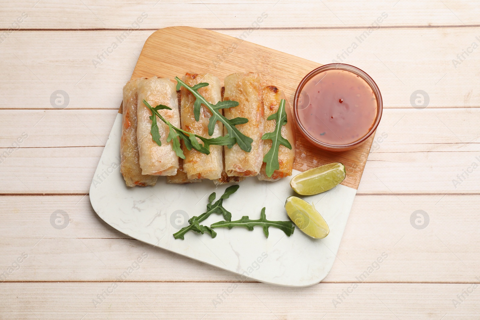 Photo of Tasty fried spring rolls, lime, arugula and sauce on light wooden table, top view