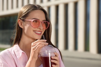 Young woman in sunglasses with plastic cup of fresh juice outdoors, space for text