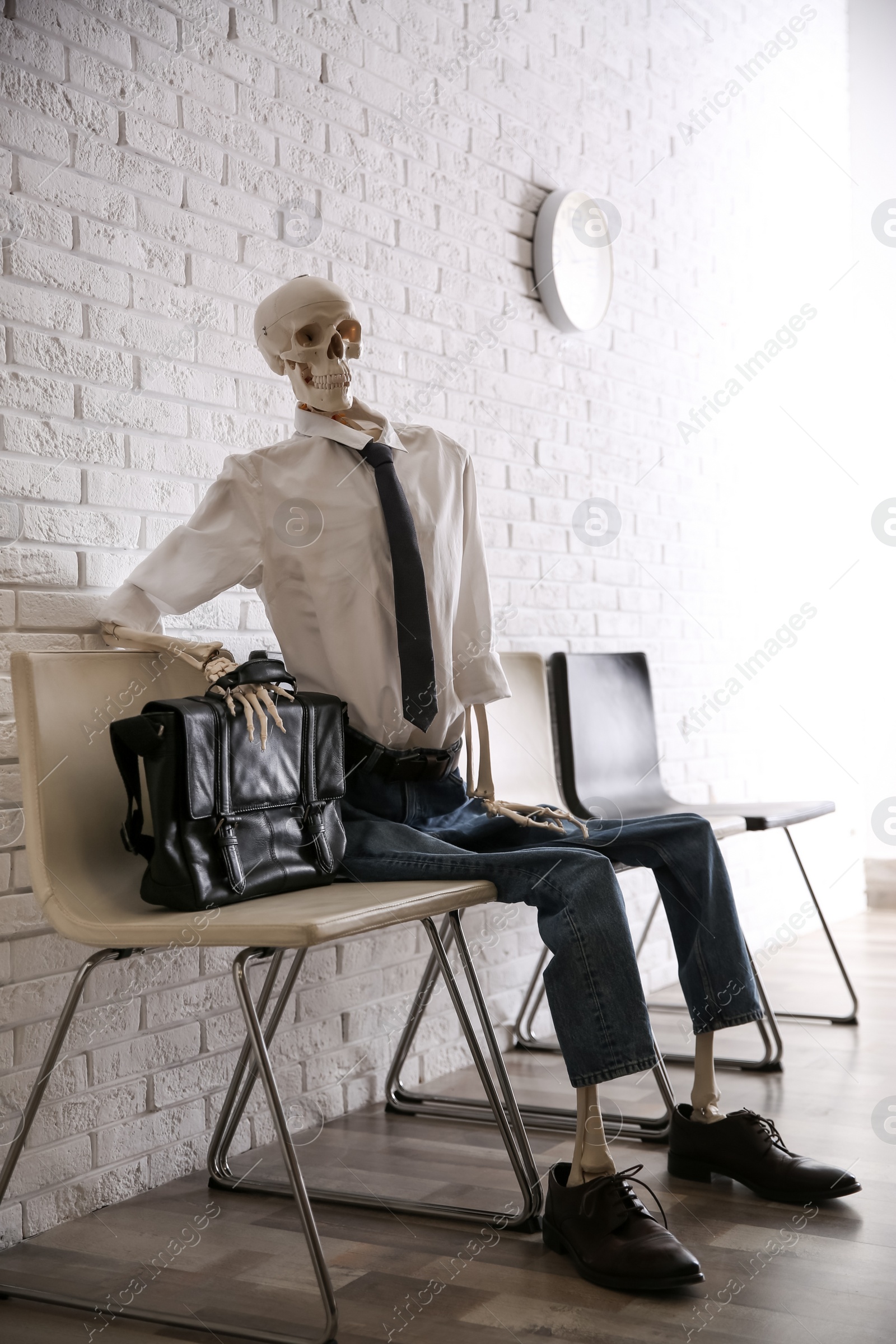 Photo of Human skeleton in office wear sitting on chair near brick wall indoors