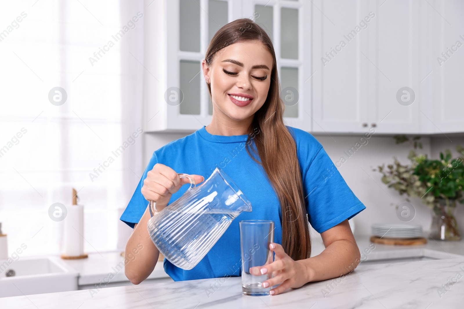 Photo of Happy woman pouring water from jug into glass at white marble in kitchen