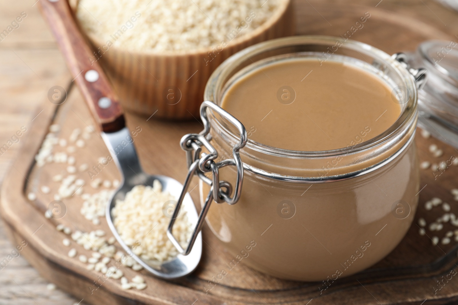 Photo of Jar of tasty sesame paste and seeds on wooden board, closeup