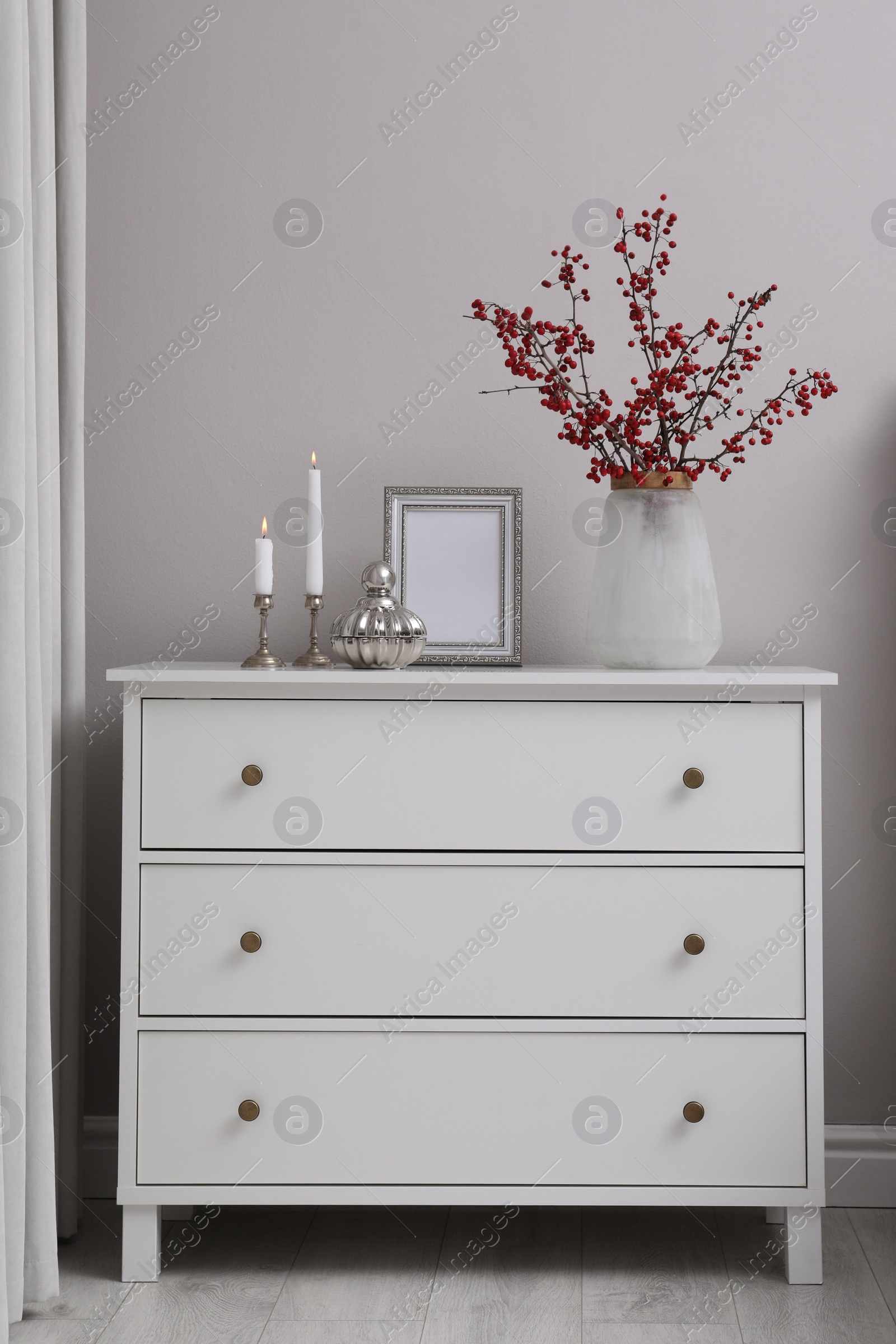 Photo of Hawthorn branches with red berries in vase, candles and frame on chest of drawers indoors