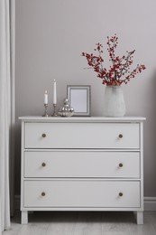 Hawthorn branches with red berries in vase, candles and frame on chest of drawers indoors