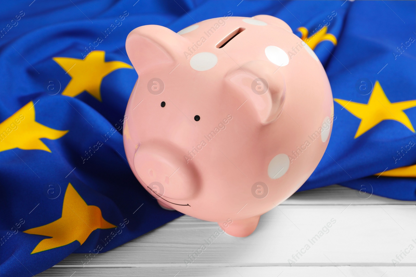 Photo of Pink piggy bank and flag of European Union on white wooden table, closeup