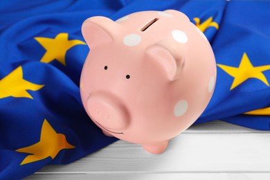 Pink piggy bank and flag of European Union on white wooden table, closeup