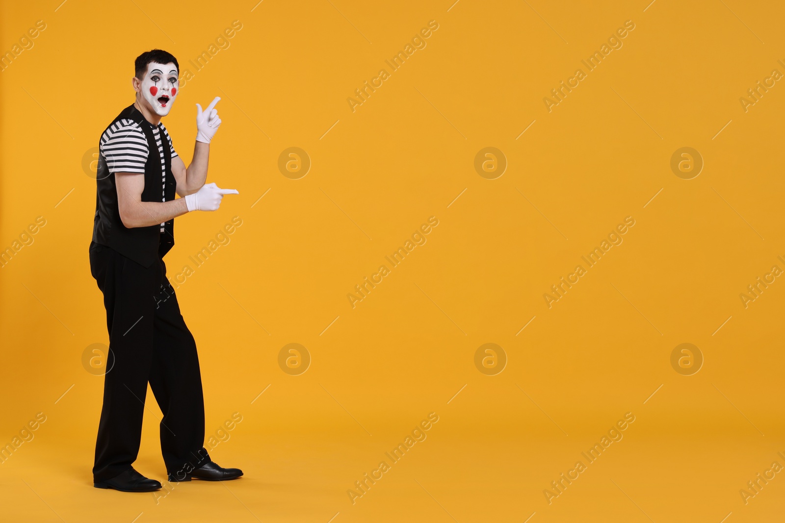 Photo of Funny mime artist pointing at something on orange background. Space for text