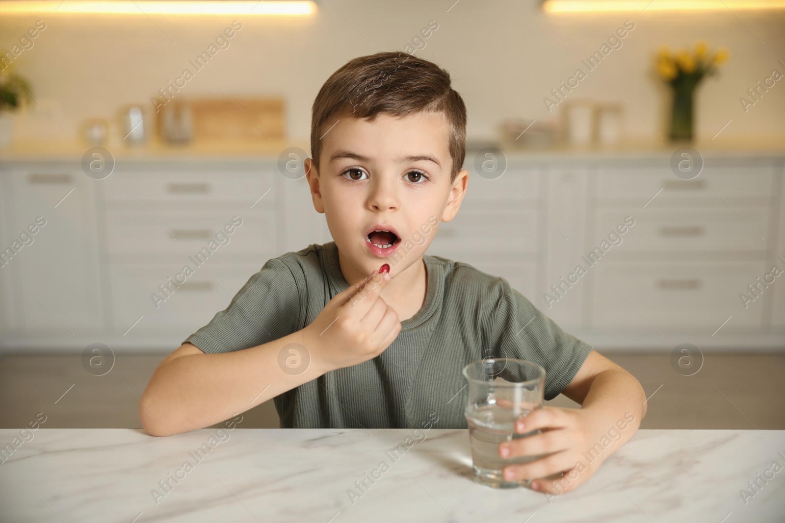 Photo of Little boy with glass of water taking vitamin capsule in kitchen