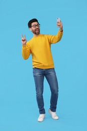 Smiling man taking selfie with smartphone and showing peace sign on light blue background