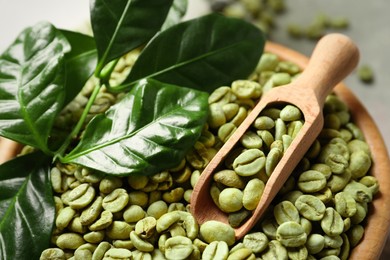 Photo of Green coffee beans, wooden scoop and leaves in bowl, closeup