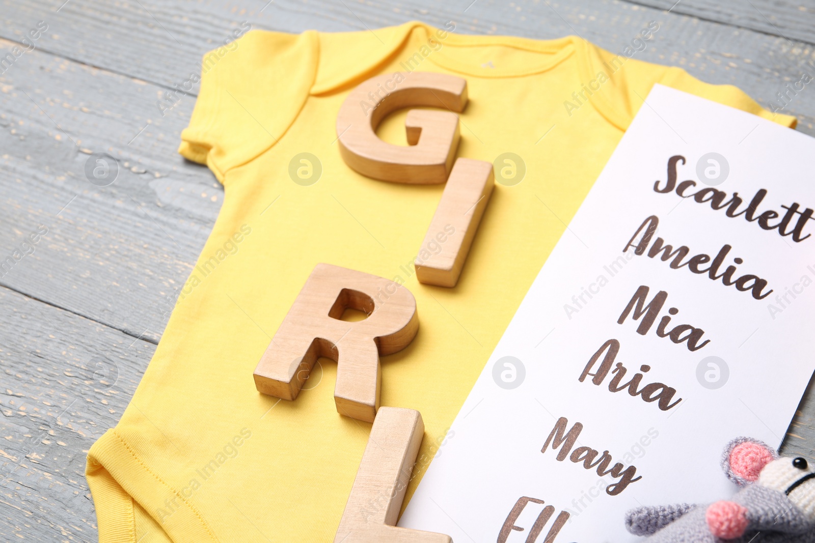 Photo of Bodysuit, list of baby names and toys on grey wooden background, closeup