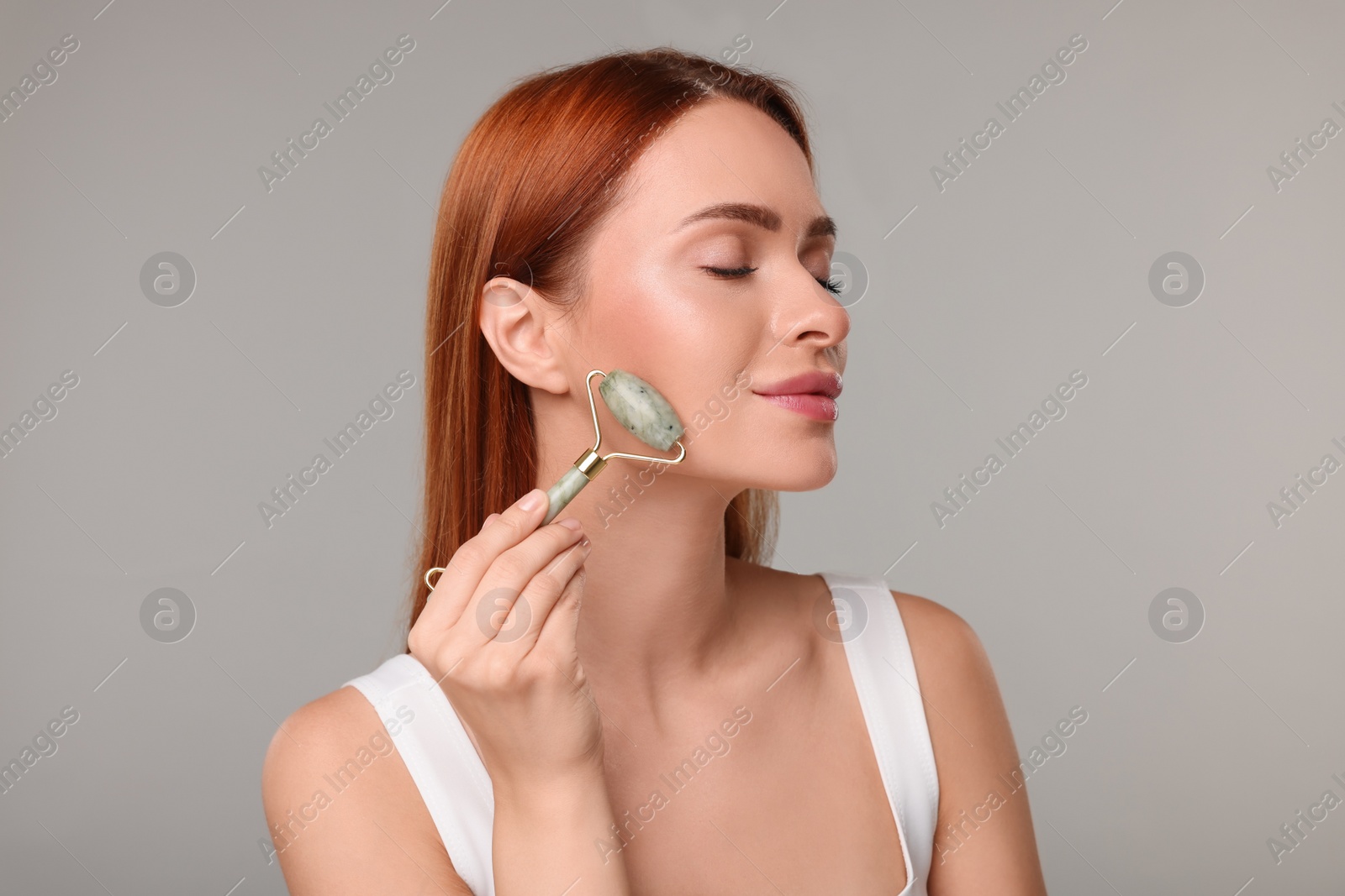 Photo of Young woman massaging her face with jade roller on grey background