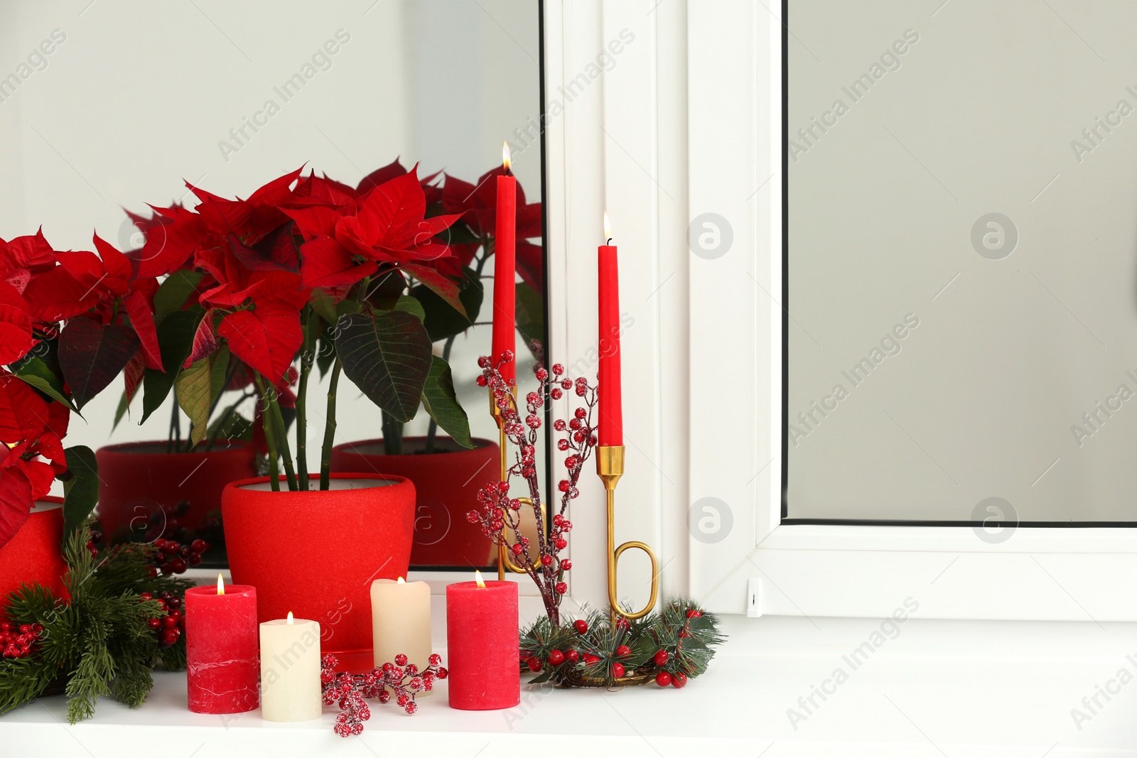 Photo of Potted poinsettias, burning candles and festive decor on windowsill in room, space for text. Christmas traditional flower