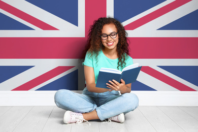 Image of Beautiful young African-American girl reading book and flag of Great Britain on wall. Learning English