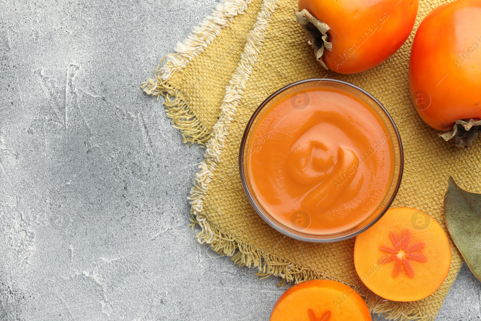 Photo of Delicious persimmon jam in glass bowl and fresh fruits on gray textured table, flat lay. Space for text