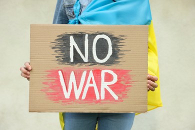 Photo of Young woman wrapped in Ukrainian flag holding poster with words No War near light wall, closeup