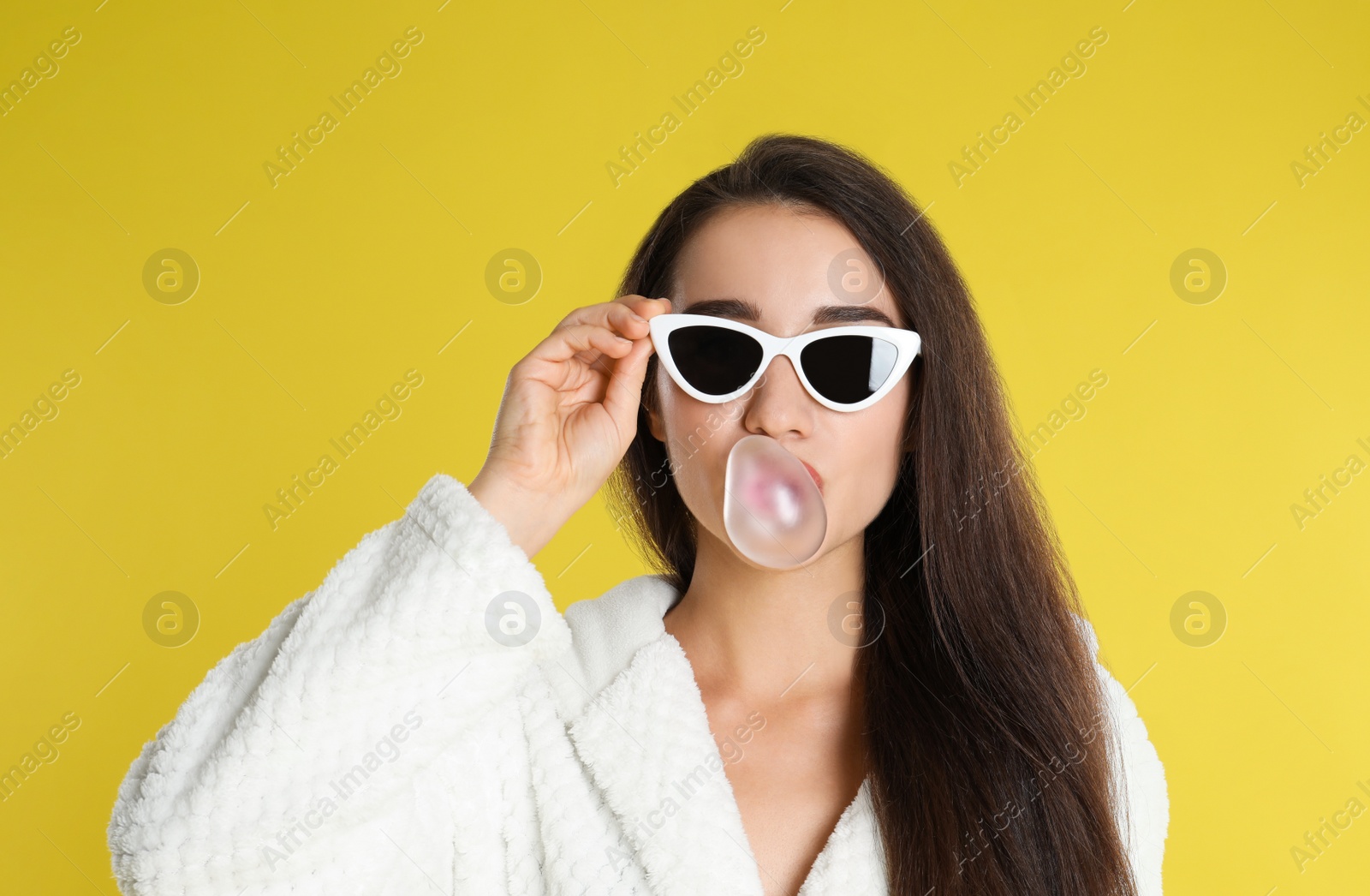 Photo of Young woman in bathrobe and sunglasses blowing chewing gum on yellow background