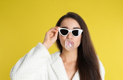 Photo of Young woman in bathrobe and sunglasses blowing chewing gum on yellow background