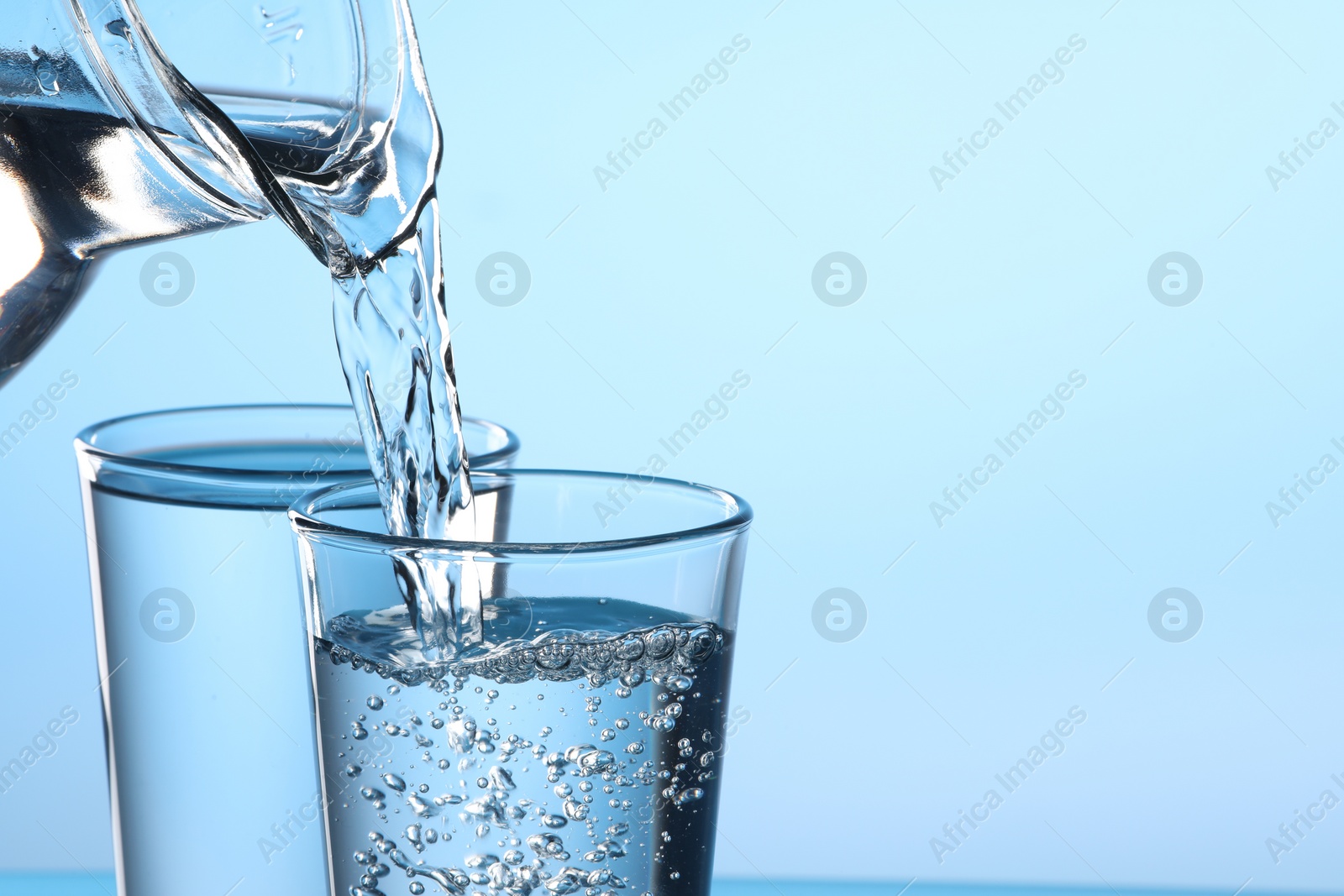 Photo of Pouring water from jug into glass on light blue background, closeup. Space for text