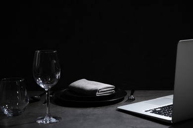 Composition with tableware and laptop on table against black background, space for text. Loneliness concept