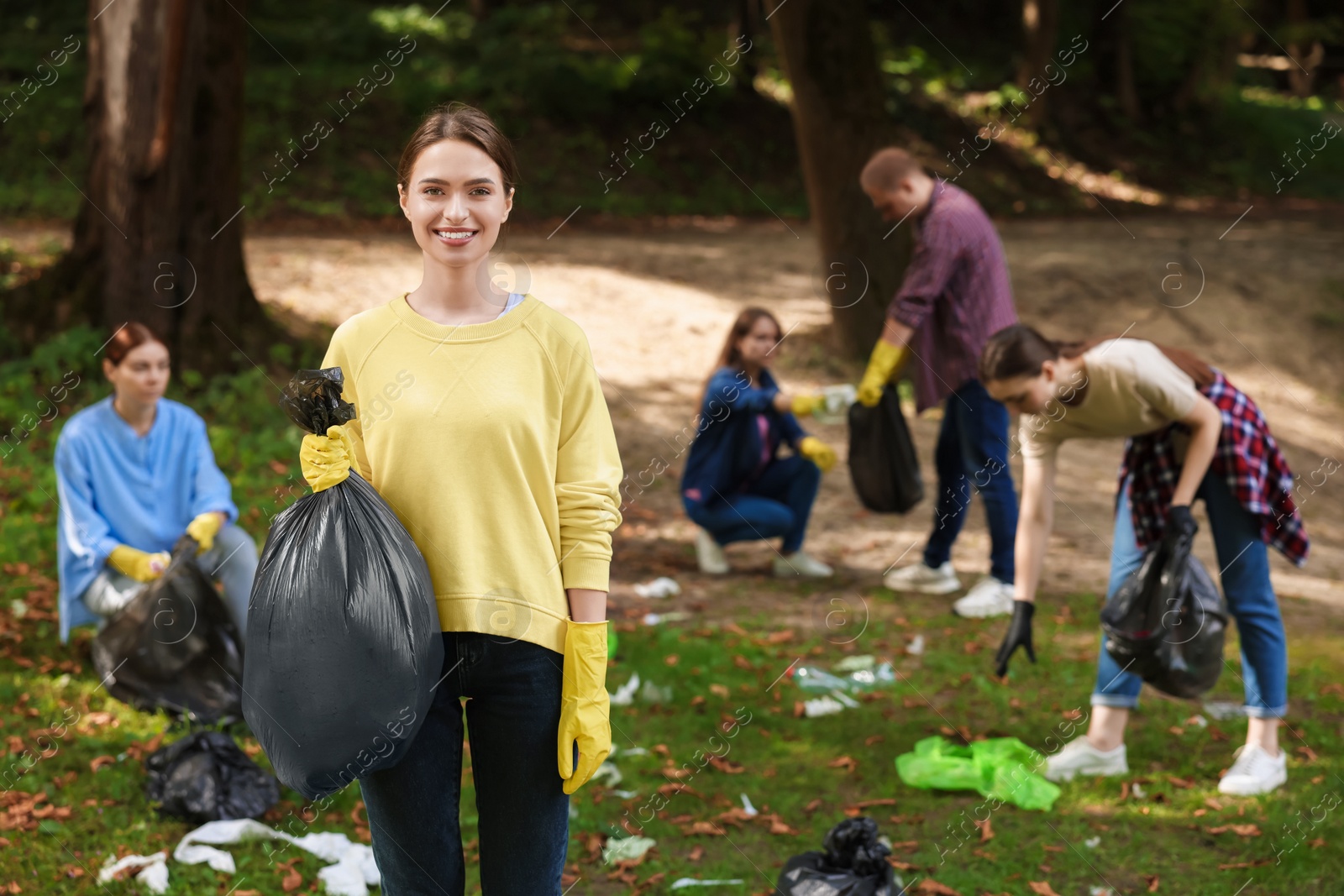 Photo of Young woman with plastic bag and group of people collecting garbage in park