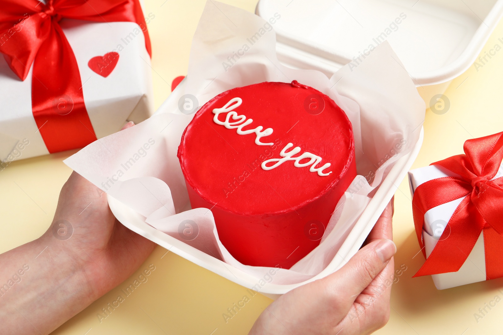 Photo of Woman holding takeaway box with bento cake at beige table, closeup. St. Valentine's day surprise