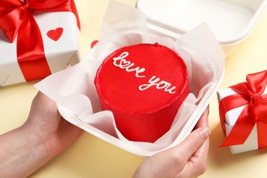 Photo of Woman holding takeaway box with bento cake at beige table, closeup. St. Valentine's day surprise